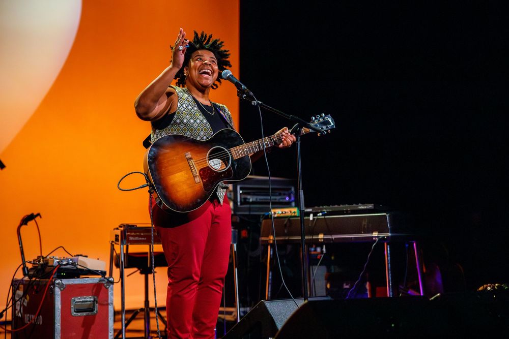 Energetic performer plays acoustic guitar on stage with vibrant orange lighting and music equipment.