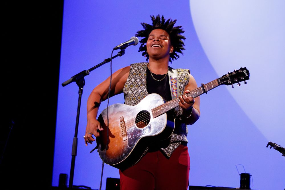 Musician performs on stage with an acoustic guitar against a blue backdrop during a live concert performance.