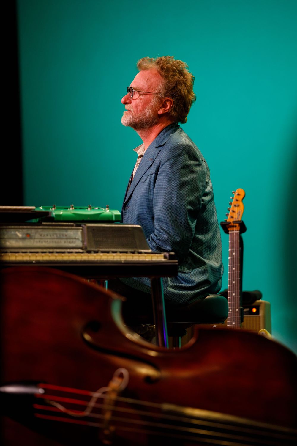 Musician in blue blazer performs at piano against turquoise backdrop with bass guitar visible in foreground.