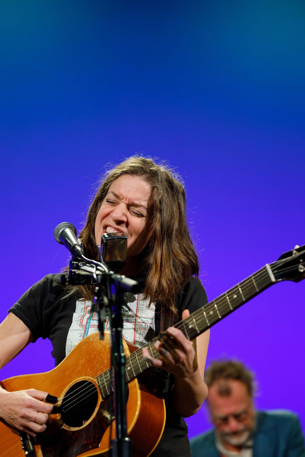 Musical performer singing passionately with acoustic guitar against blue gradient backdrop.