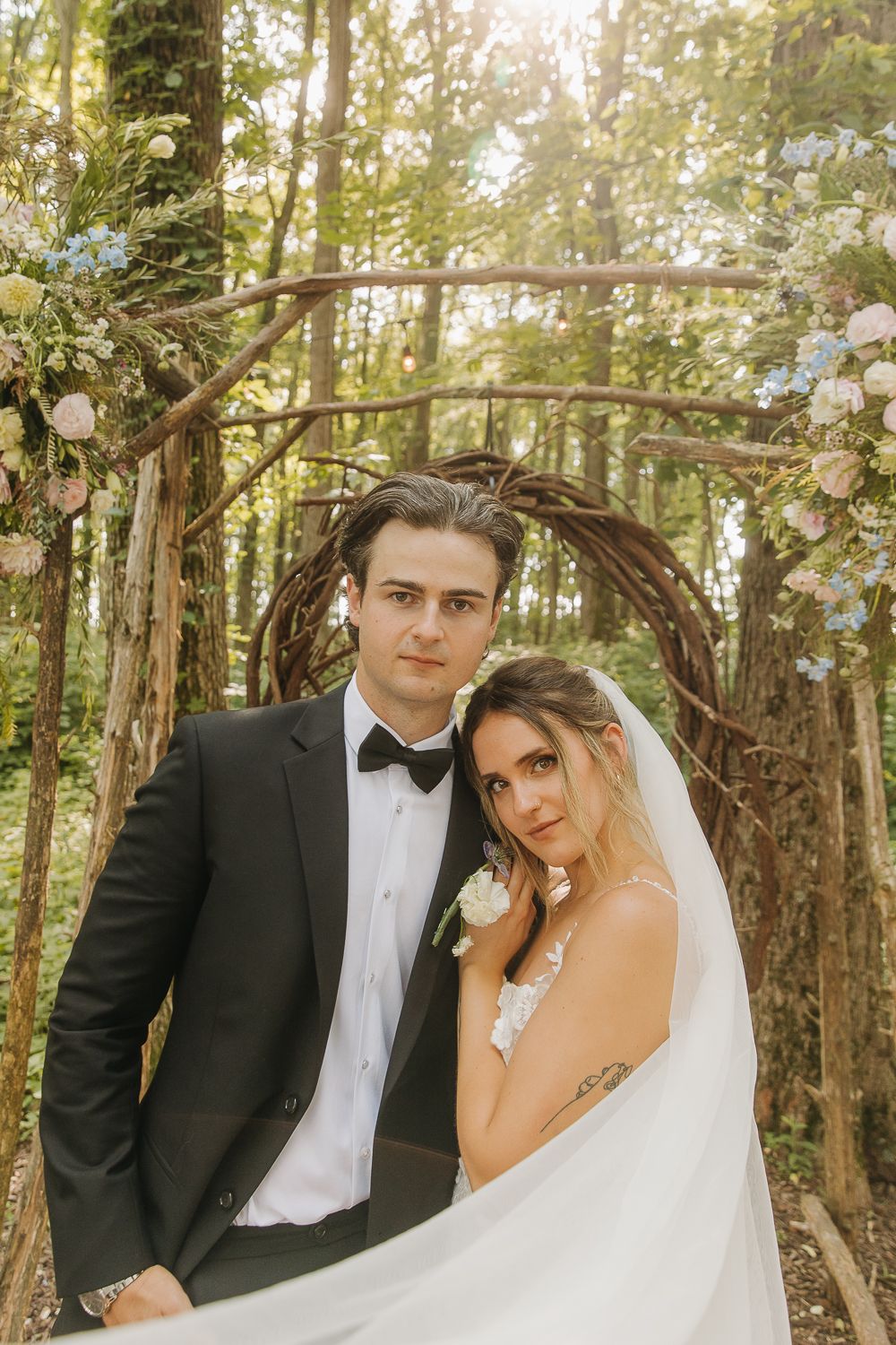 A wedding couple poses under a rustic wooden arch decorated with white flowers in a sunlit forest setting.