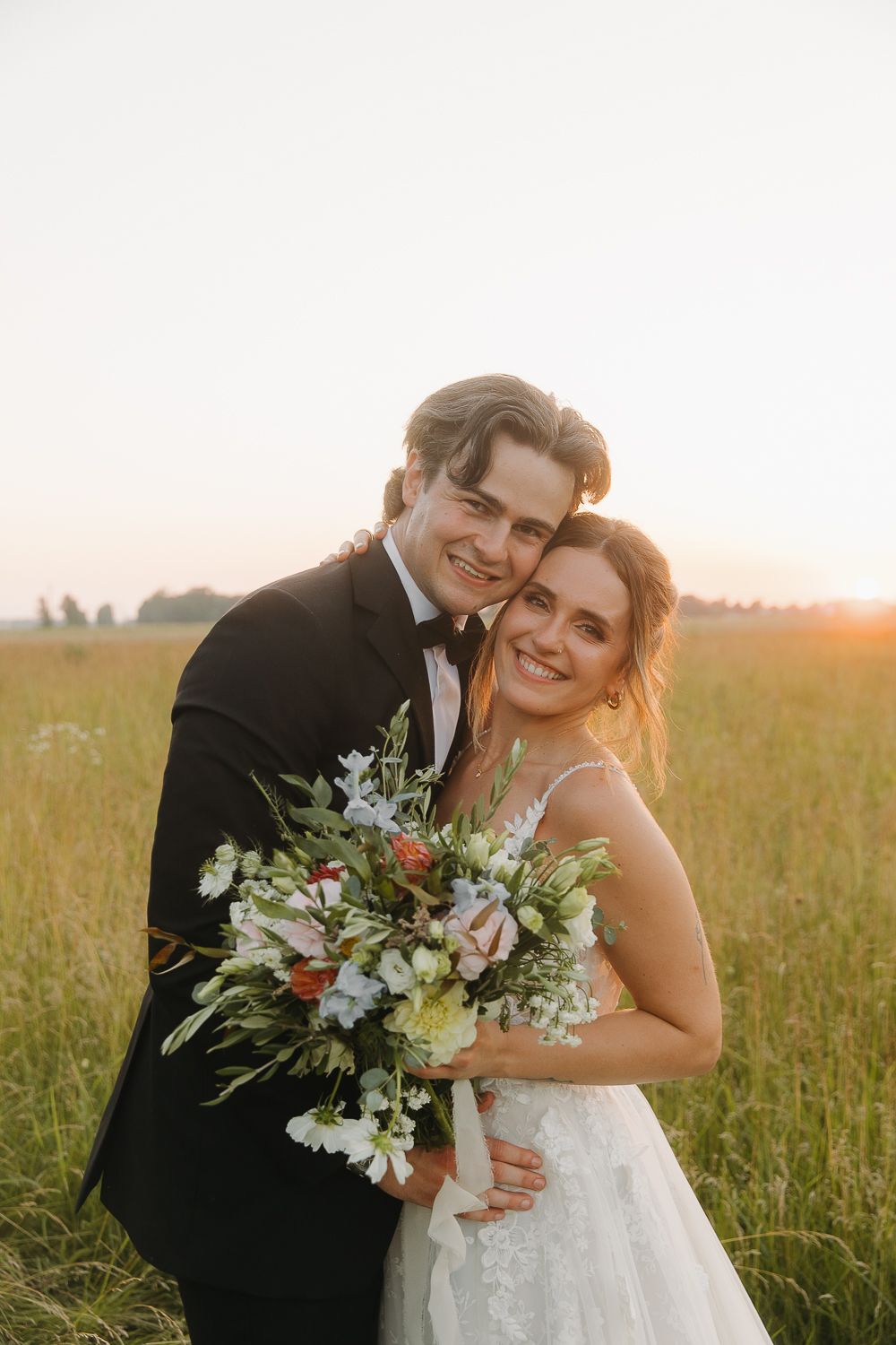 A happy wedding couple embraces in a golden meadow during sunset while holding a beautiful wildflower bouquet.