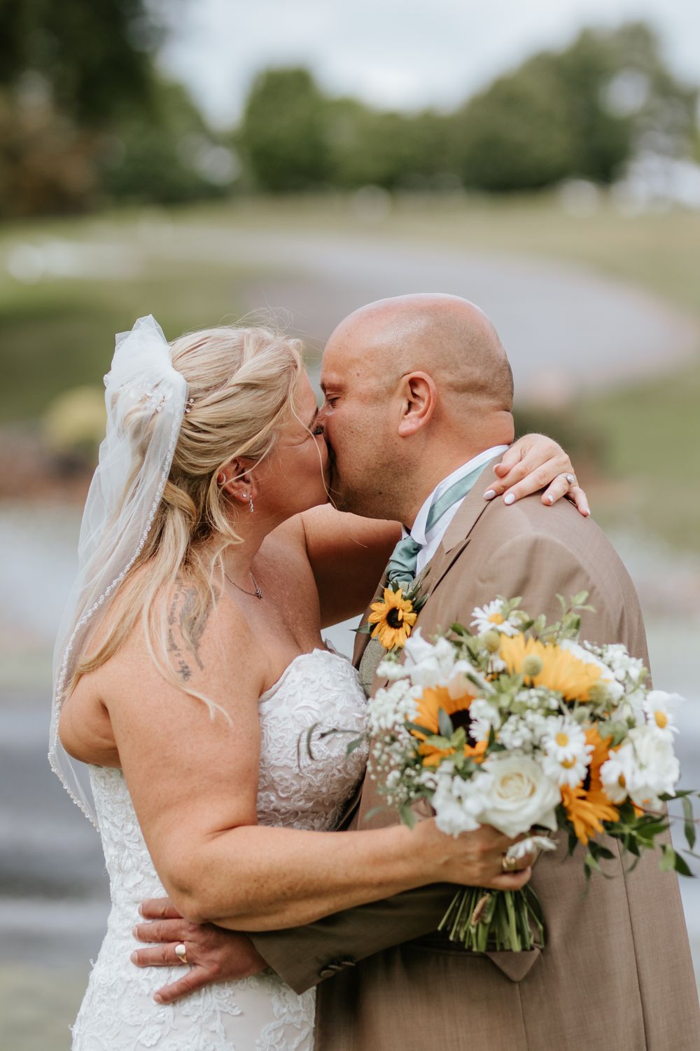 A tender wedding kiss between newlyweds captured outdoors, with the bride holding a beautiful sunflower and white flower bouquet.
