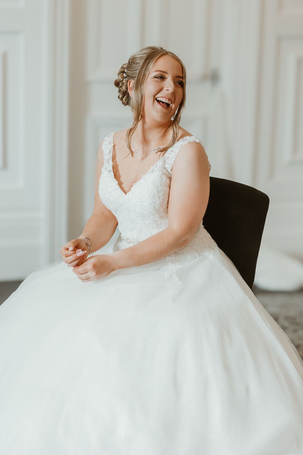 A joyful bride wearing a white lace ballgown wedding dress laughs while sitting in a chair. Somerset and Devon Wedding Photographer