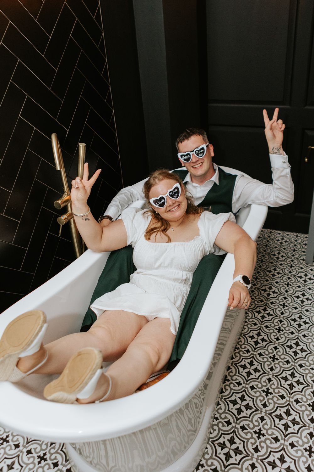 A playful couple wearing matching sunglasses pose making peace signs while sitting in a vintage white bathtub.