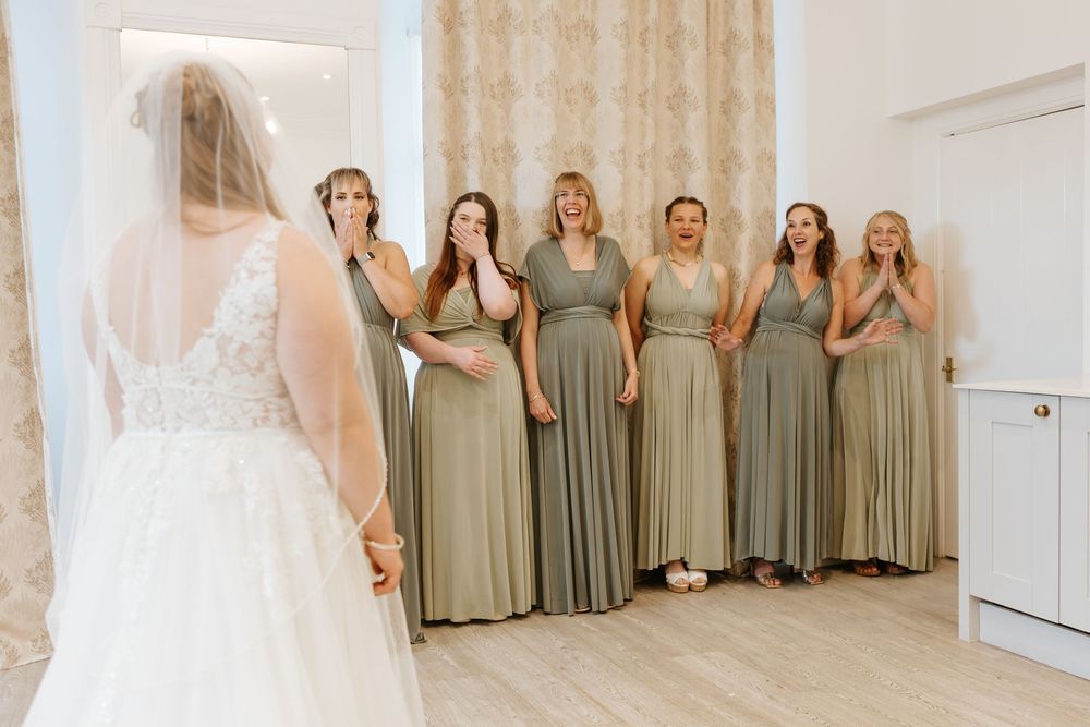 Bridesmaids in sage green dresses react with joy seeing bride in her wedding gown in sunlit room.