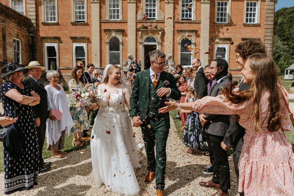 Wedding guests throw confetti at a couple outside a grand Georgian brick mansion on a sunny day.