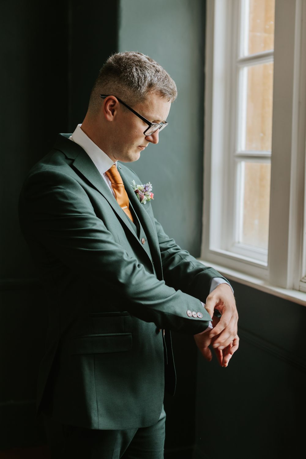 A person in a dark green suit and orange tie stands by window in moody lighting creating an atmospheric scene.