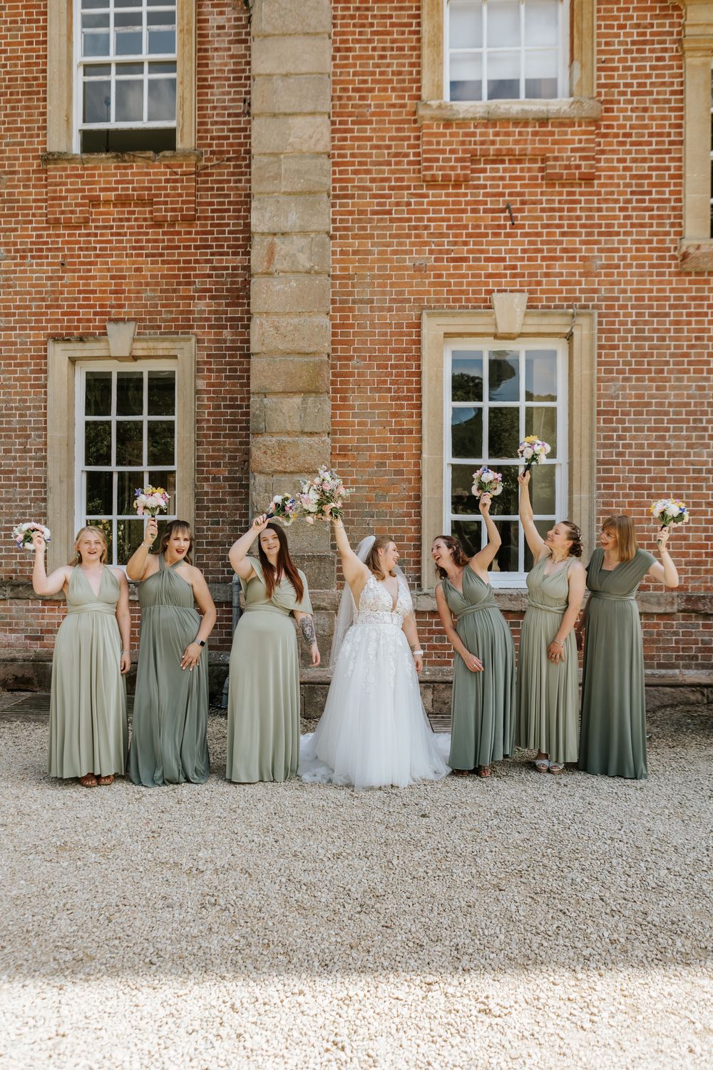 Wedding party poses together in sage green bridesmaid dresses and white bridal gown against historic brick building.