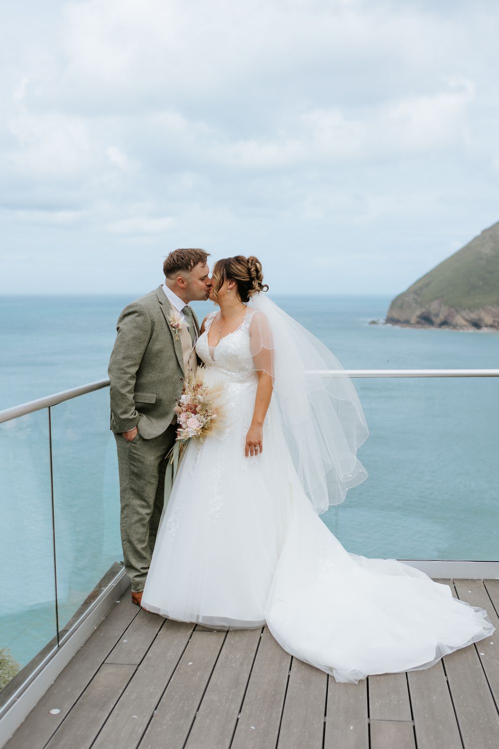 A bride and groom share an intimate moment on a coastal deck overlooking blue ocean waters and mountains.