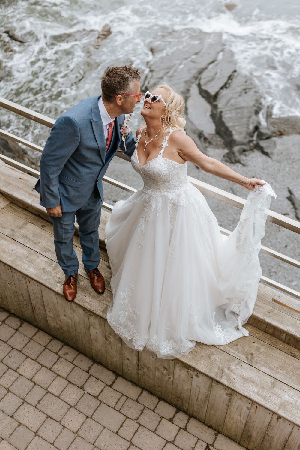A romantic wedding couple sitting together on a stone ledge while sharing intimate moments in their formal attire.