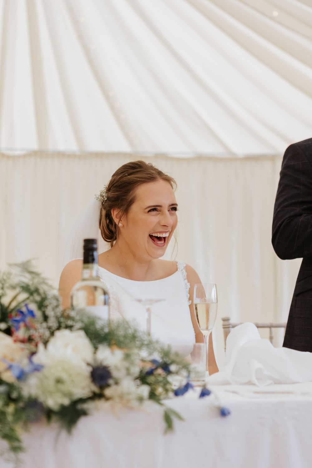 A joyful bride laughs during wedding reception speeches beside elegant floral centerpieces in a white tent.