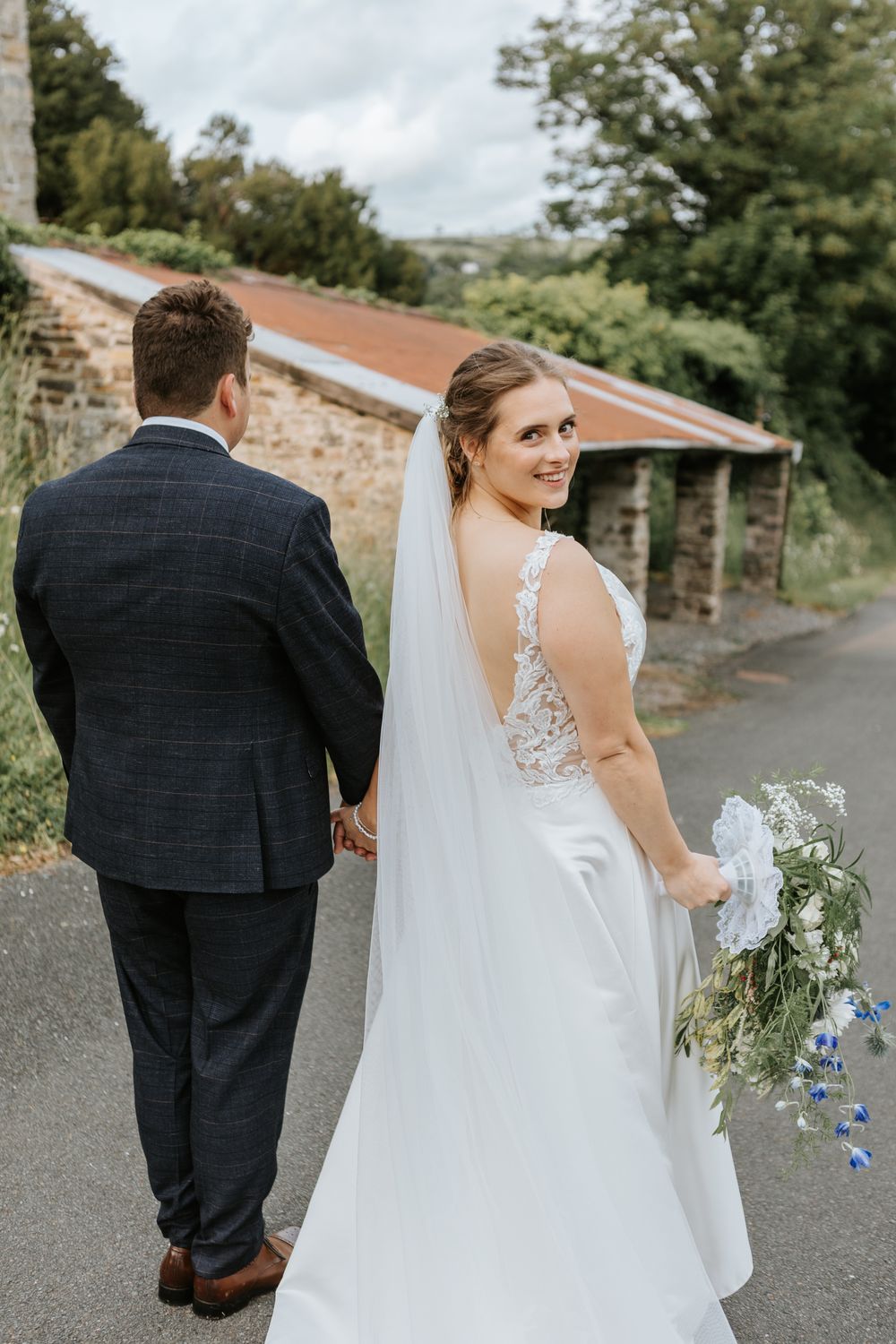 Bride in elegant white lace dress with flowing veil holds bouquet while walking with groom in navy suit along stone path.