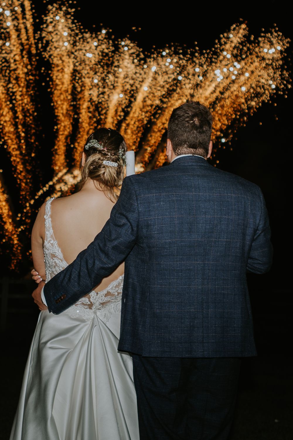 Newlywed couple watches fireworks display together while wearing wedding attire at night celebration.