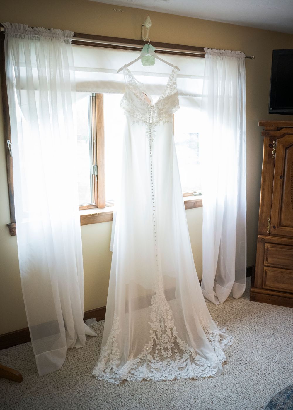 A beautiful white lace wedding dress hangs by a window, illuminated by natural light streaming through sheer curtains.