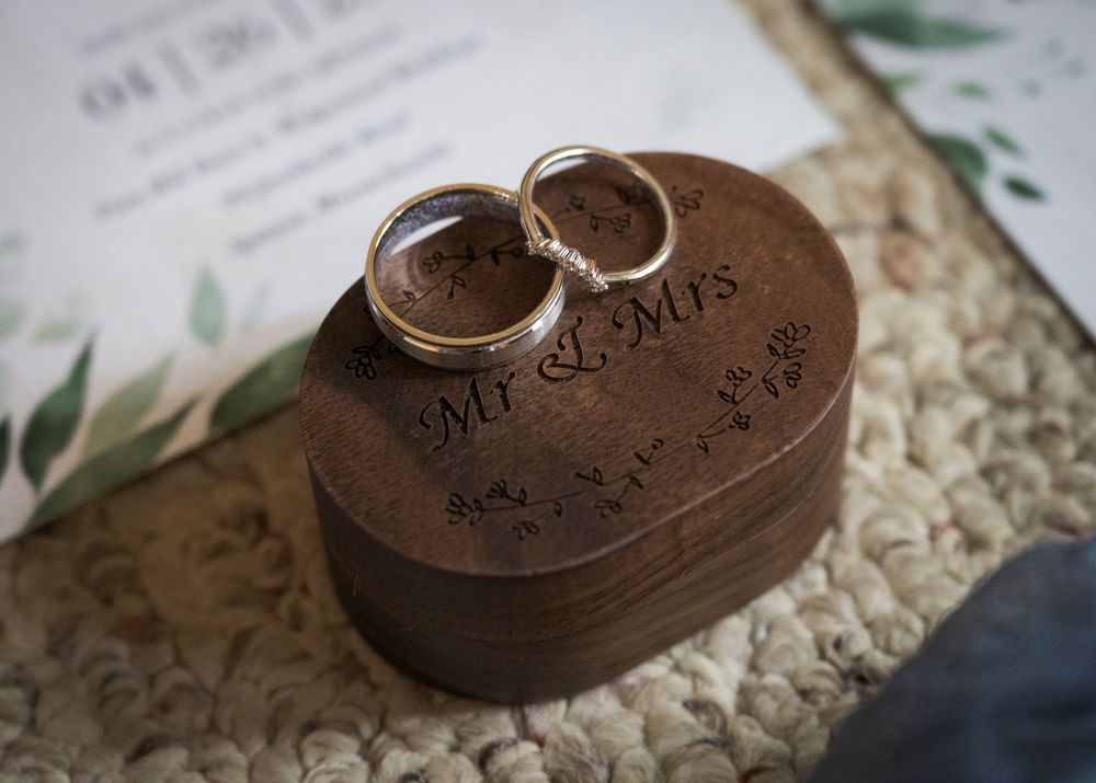 Two wedding rings sit atop a wooden heart-shaped ring box with engraved text and botanical elements in the background.