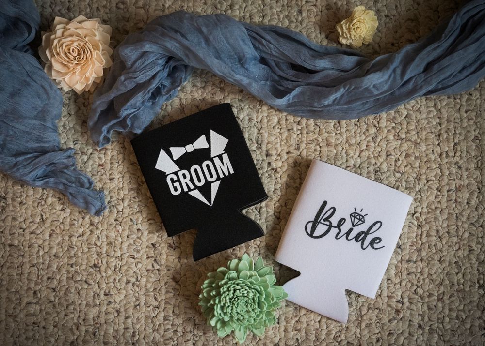Wedding accessories featuring bride and groom drink koozies with a blue fabric and floral decorations on burlap.