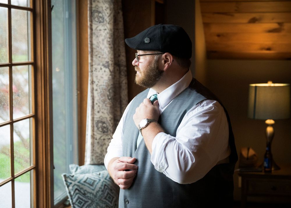 A person adjusts their tie while standing by a window in formal wedding attire.