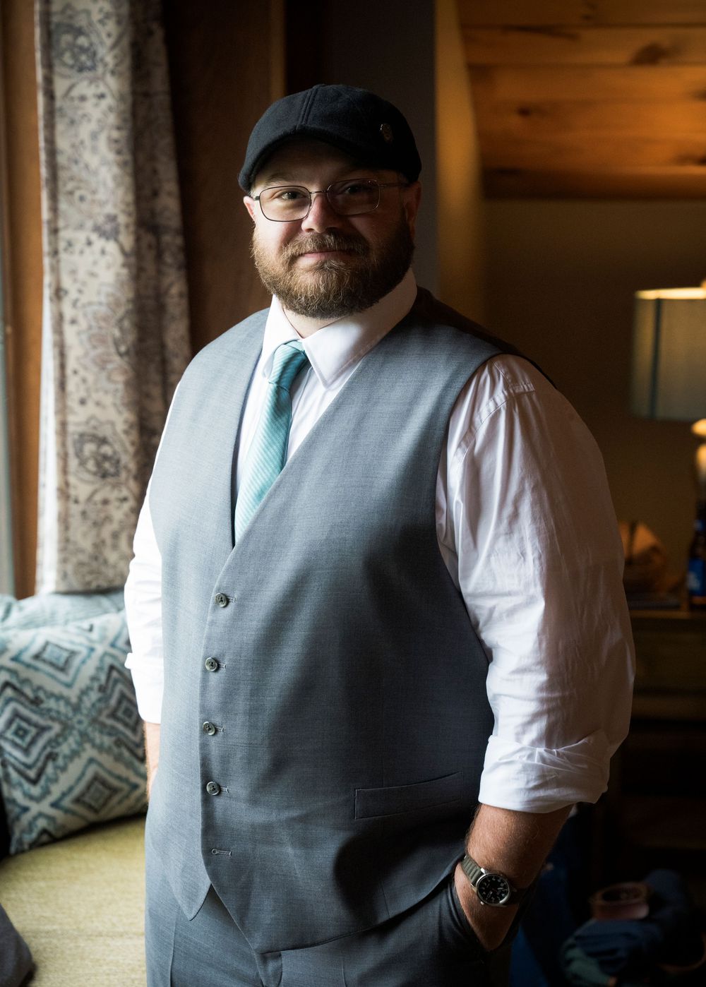 A person wearing a gray vest, white dress shirt, and teal tie poses near a window.