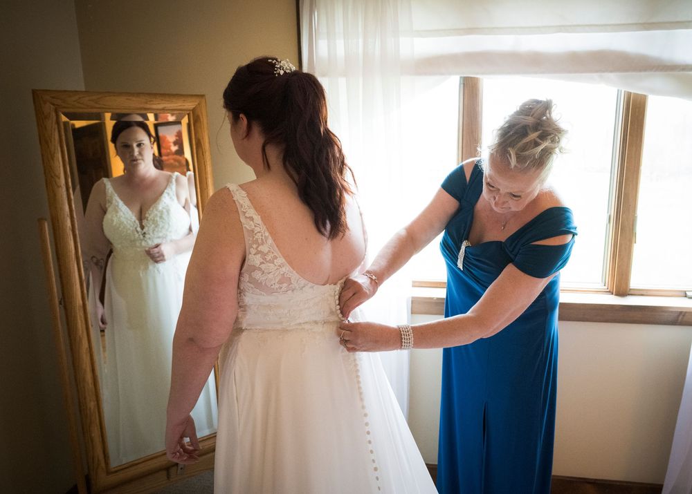 Final wedding dress preparations with helpers in a sunlit room with a full-length mirror.