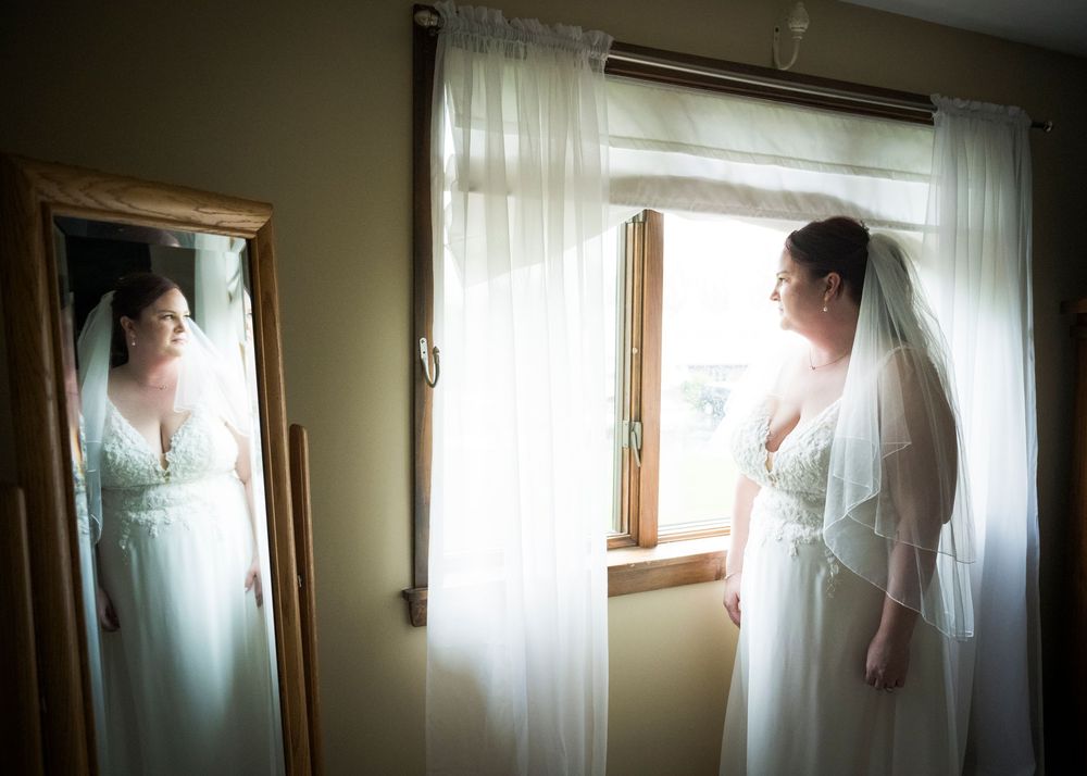 Figure in a white wedding gown standing by a window with soft natural light and a mirror reflection.