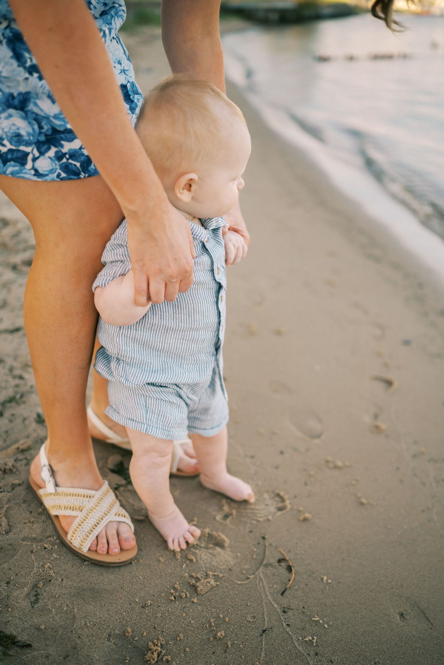 Baby taking first steps on beach sand while supported by adult in blue floral dress and sandals.