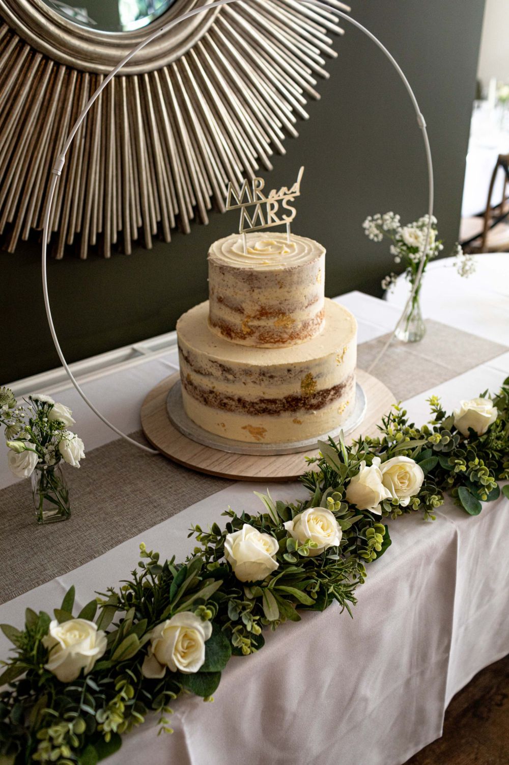 A rustic semi-naked wedding cake displayed on a table with white roses and greenery garland beneath a sunburst mirror.