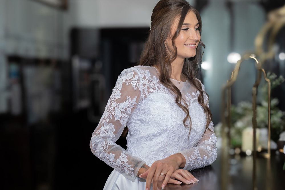 A bride wearing an elegant white lace wedding dress with long sleeves smiles while standing in a dimly lit room.