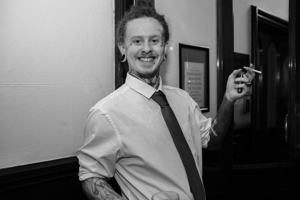 A person in a white dress shirt and black tie smiles while holding a drink in their hand.