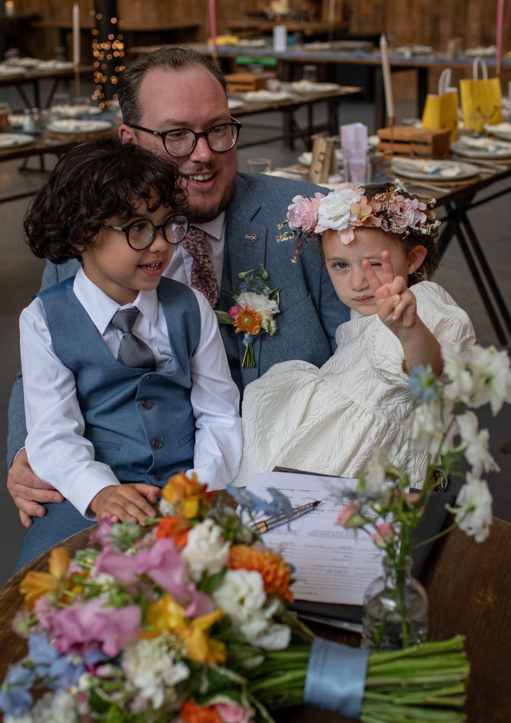 Three people share a joyful moment at a wedding reception surrounded by colorful floral arrangements and table settings.