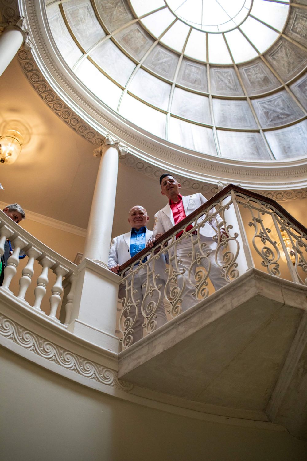 Looking up at an elegant interior staircase with decorative railings and a domed glass skylight ceiling in a historic building.