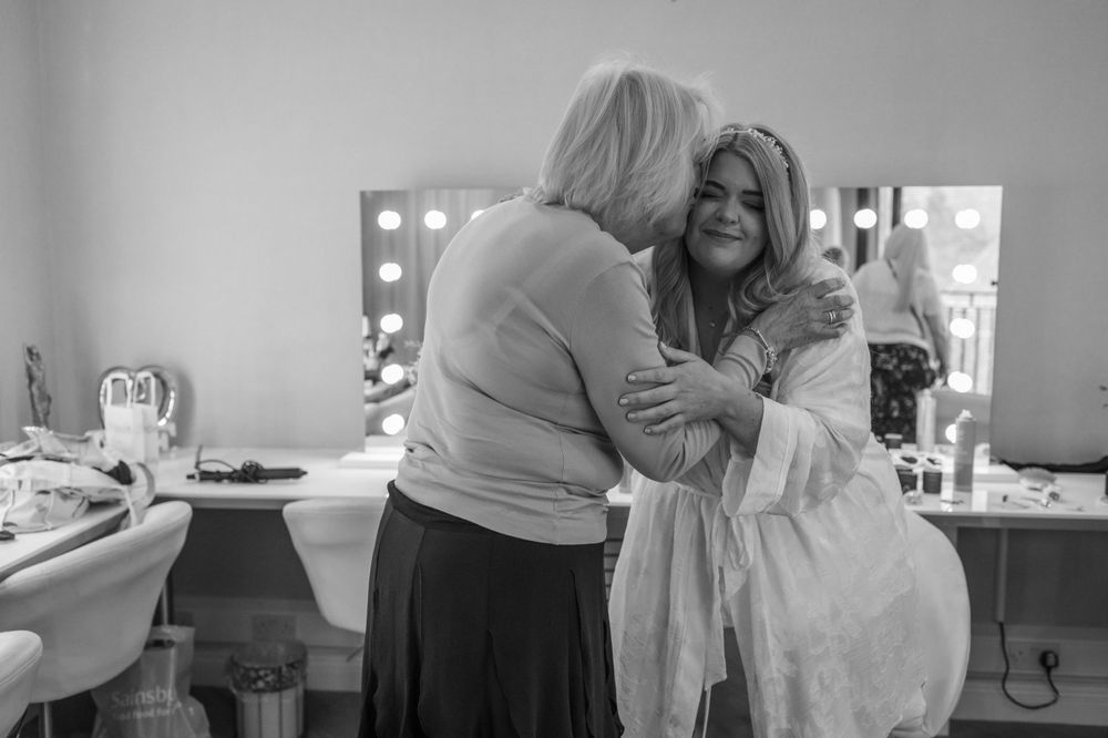 Black and white photo showing two people embracing in a makeup room with vanity mirrors.
