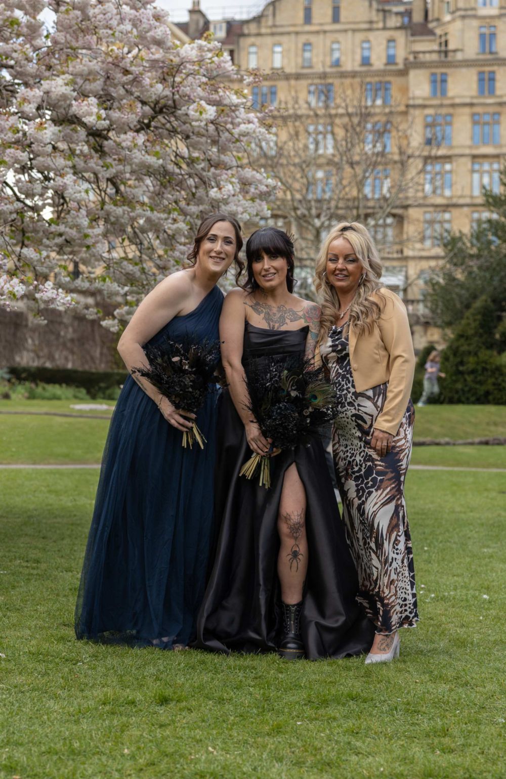 Three elegantly dressed friends pose in formal gowns beneath blooming cherry blossoms near a historic stone building.