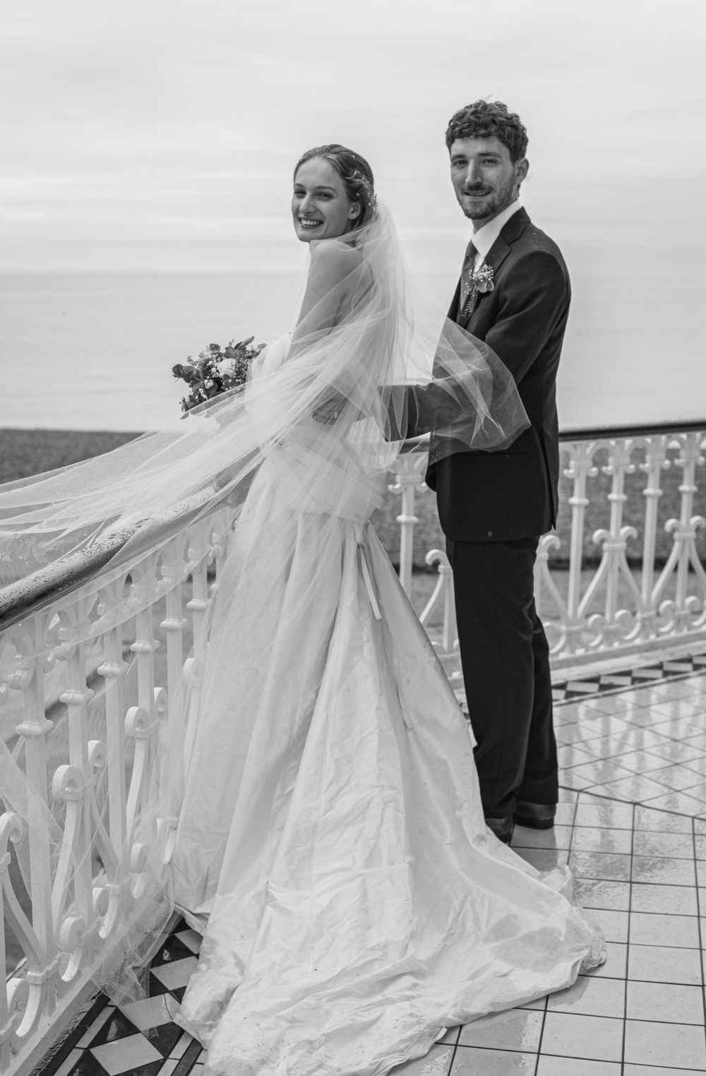 Black and white wedding photo of a couple at a seaside balcony with bride's flowing veil caught in ocean breeze.