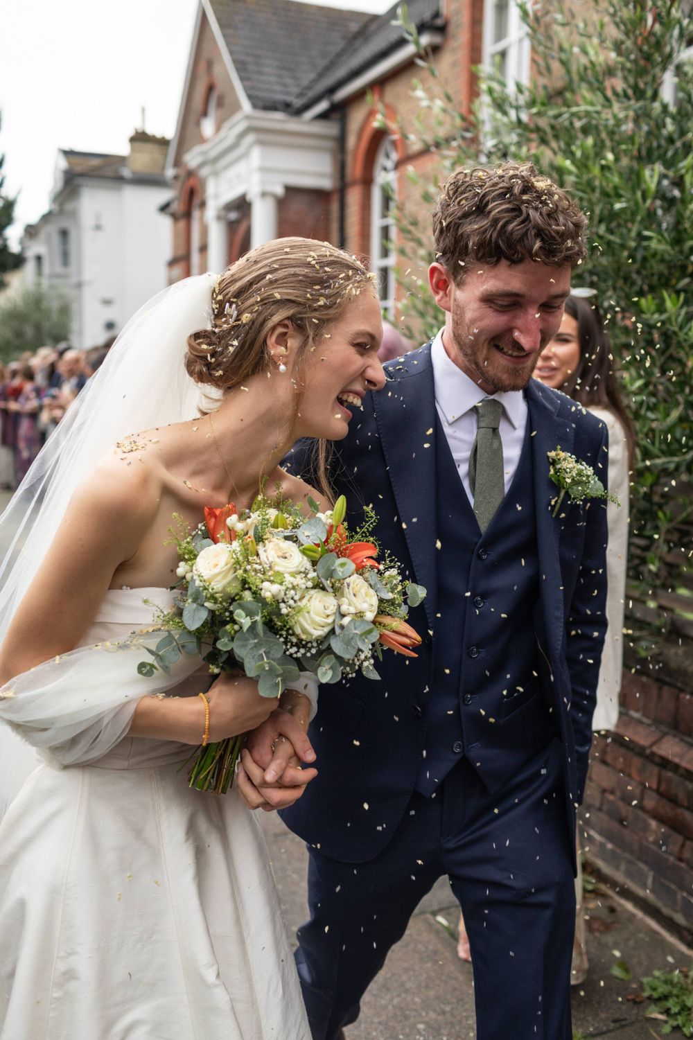 A joyful wedding couple laughing together as confetti falls around them, with the bride holding a beautiful floral bouquet.