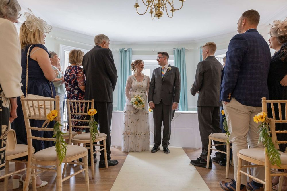 An elegant indoor wedding ceremony takes place in a bright room with gold chairs and decorative floral touches.