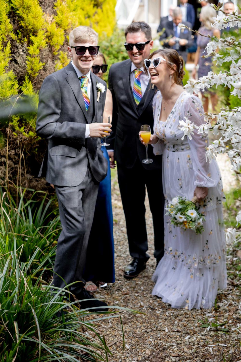 Three wedding guests share drinks and laughter on a garden path surrounded by spring blossoms and greenery.
