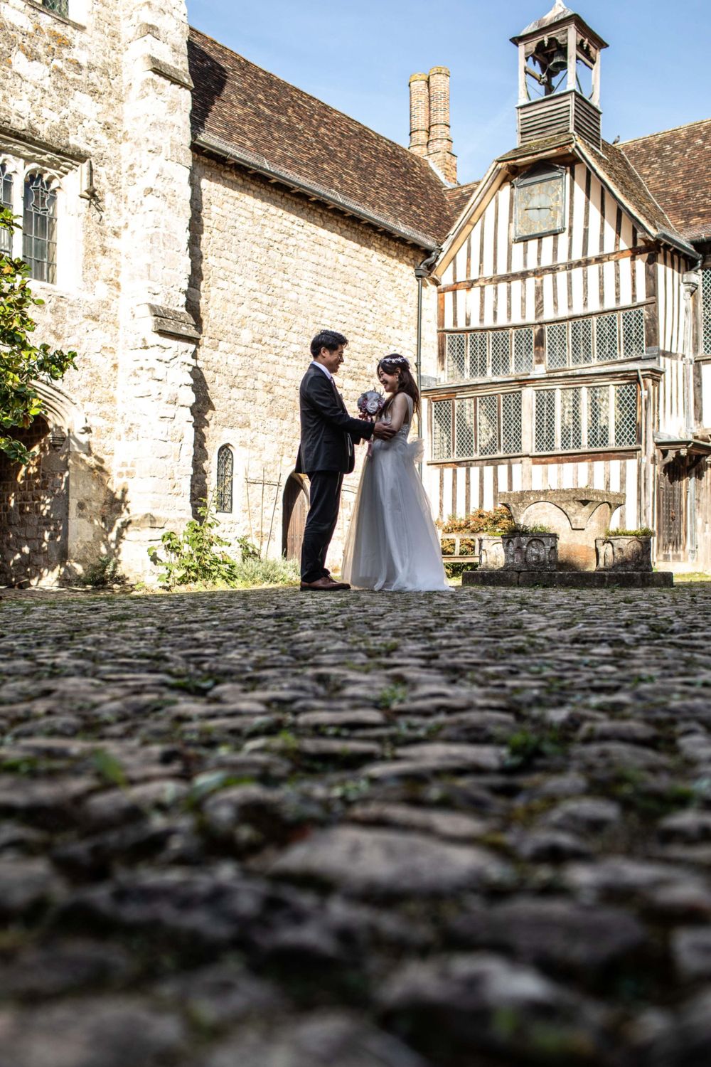 A couple shares a romantic wedding moment on cobblestone ground in front of a historic Tudor-style building with a bell tower.