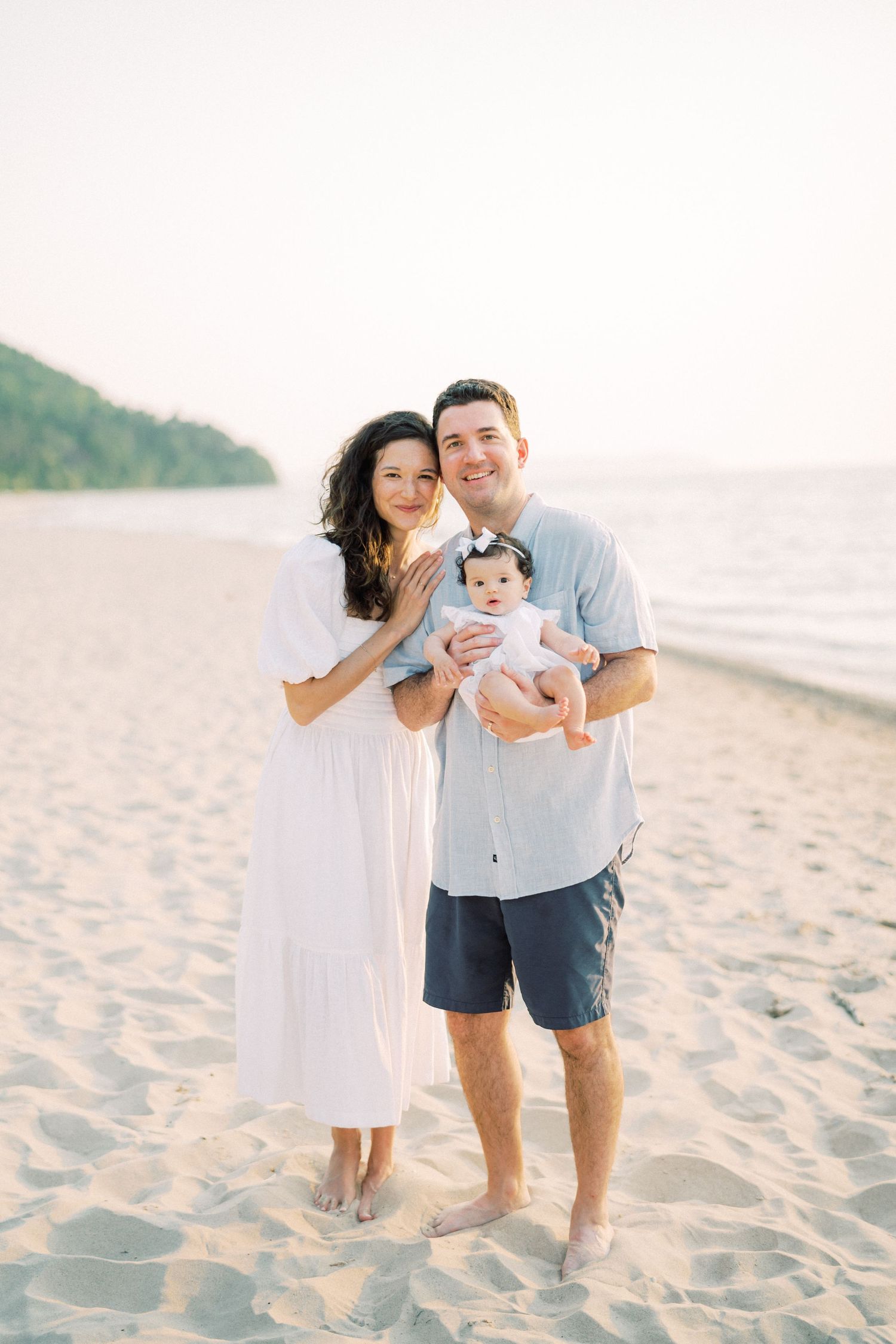 Young family poses together on sunny beach with baby in casual summer attire.