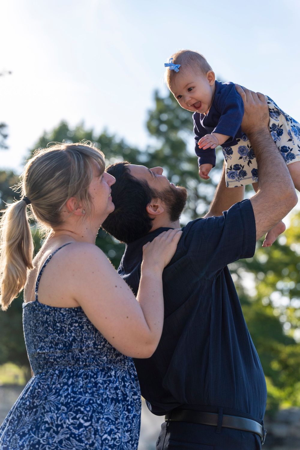 Young family shares a tender moment outdoors as they lift their baby in a floral dress.