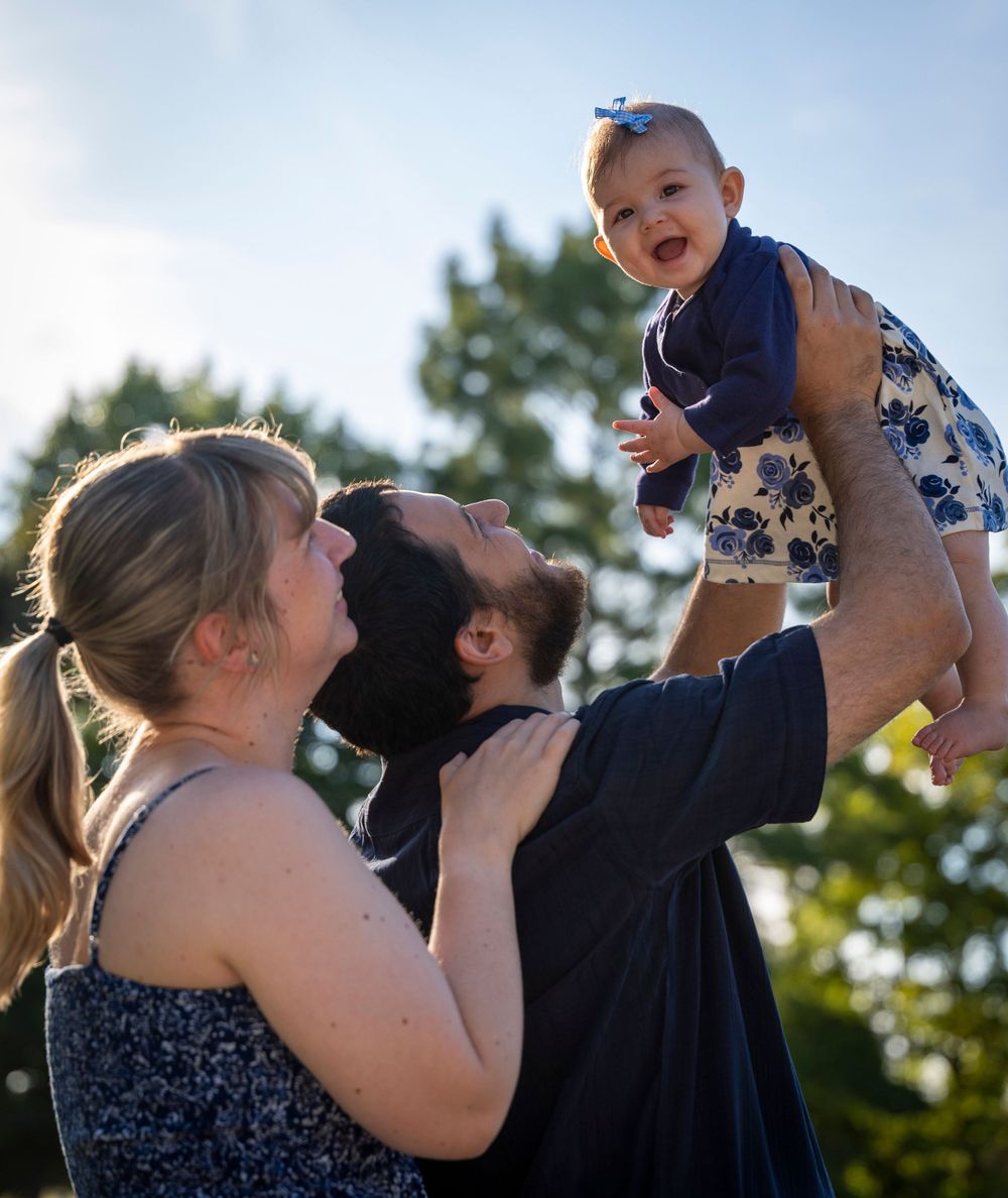 Candid outdoor family photo capturing laughter and joy as baby is lifted overhead.