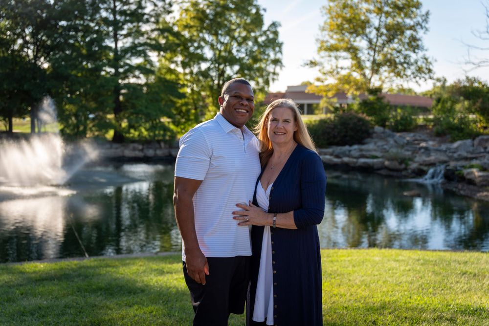 Couple stands together on grass with water fountain and pond in background.