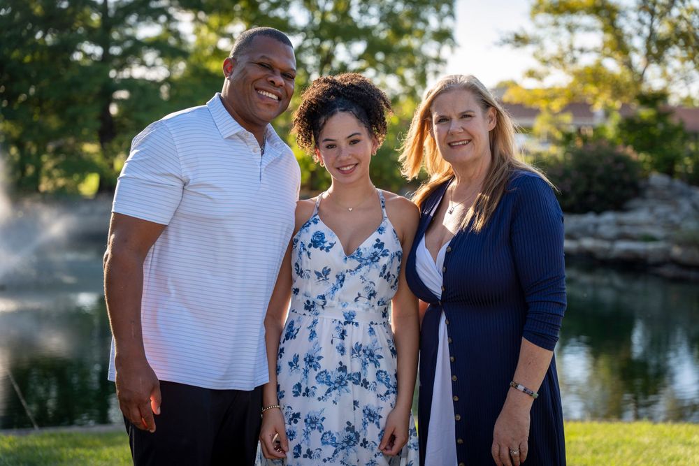 Three people pose together outdoors near a peaceful pond on a sunny day.