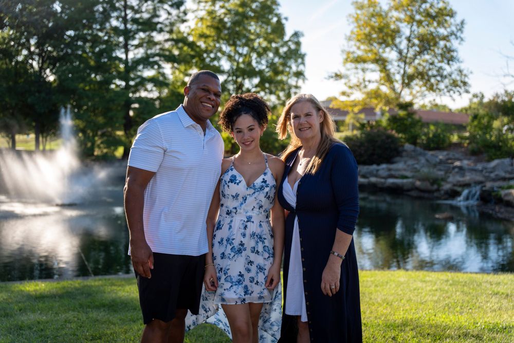 Three people smile together in front of a scenic pond with fountain spray visible.