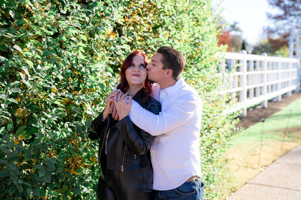 A romantic couple embraces near a white fence and green foliage on a sunny day.