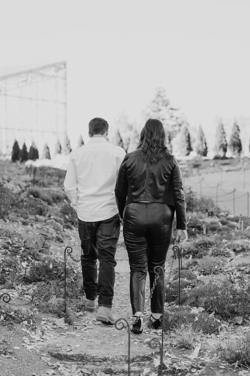 A black and white photo of a couple walking along a path with trees in the background.