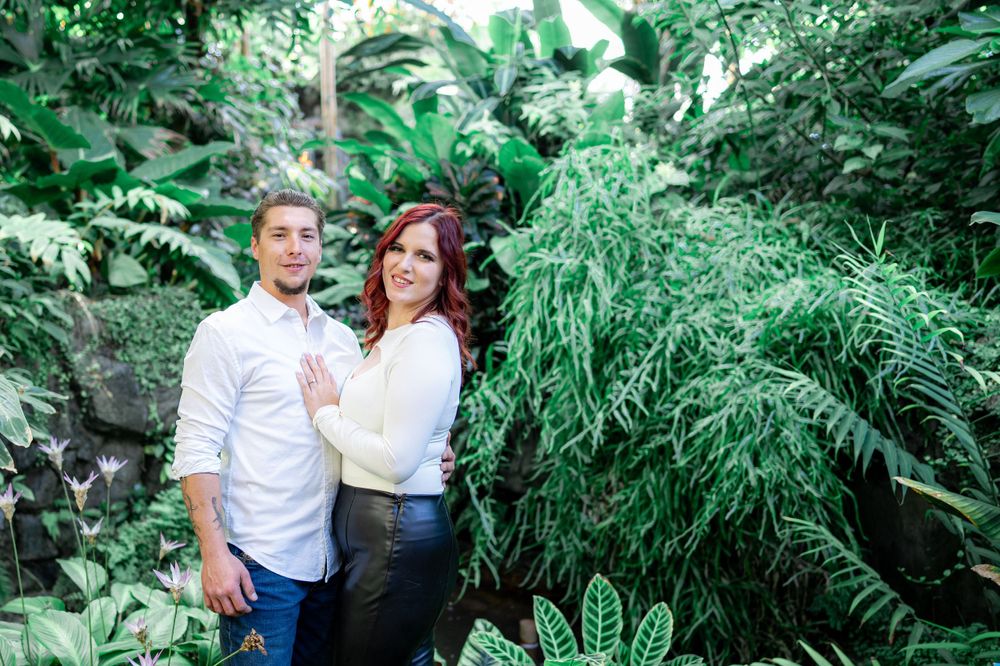 A couple poses together against lush tropical greenery in casual attire.