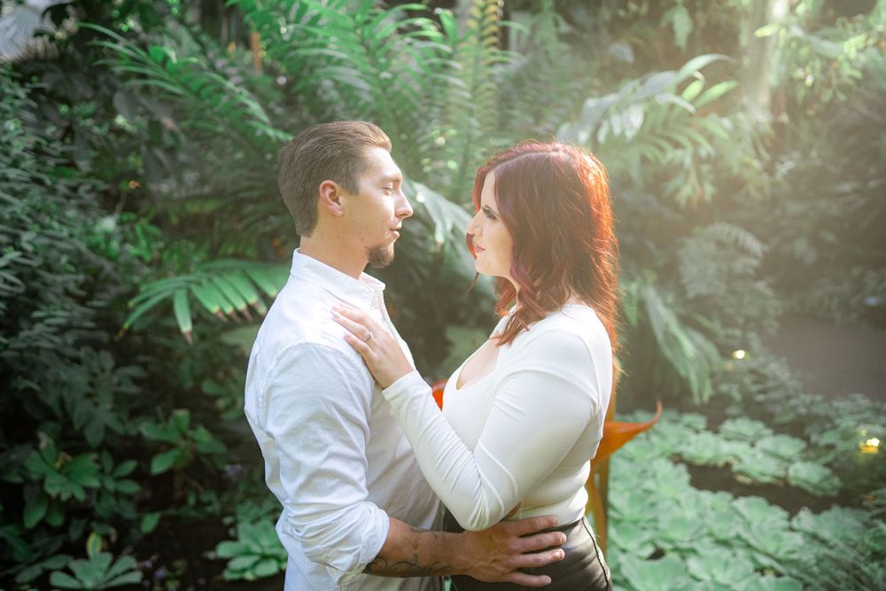 Couple embraces in a romantic garden setting with lush greenery and soft sunlight filtering through.