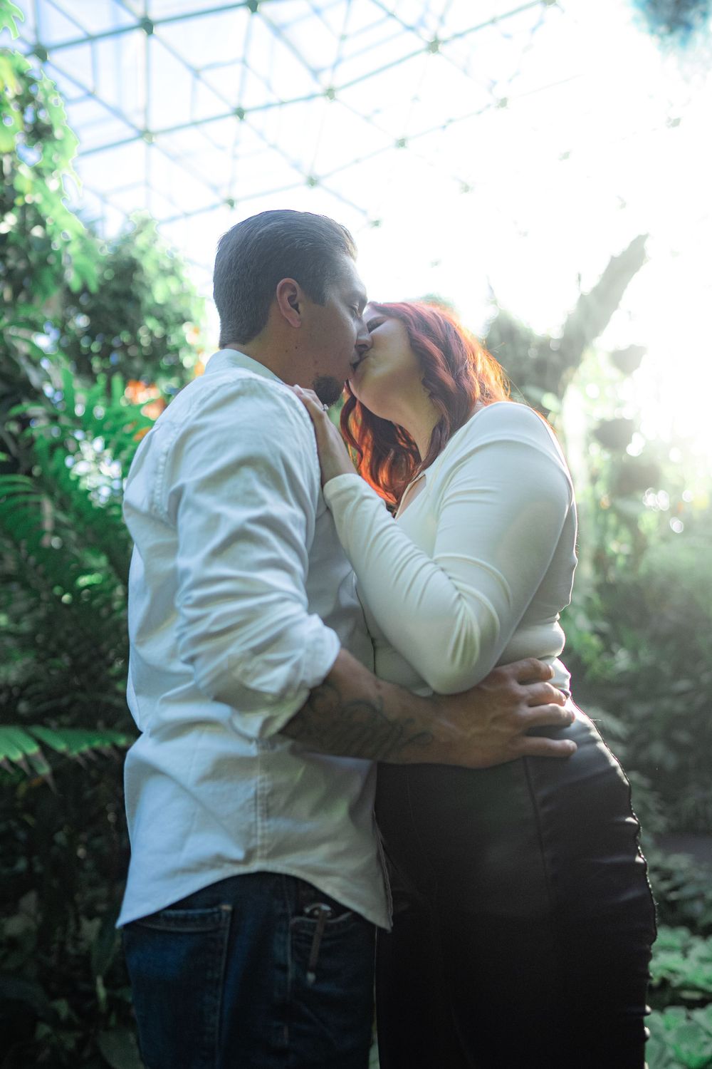 Intimate moment between couple in white shirts inside a greenhouse with natural lighting.