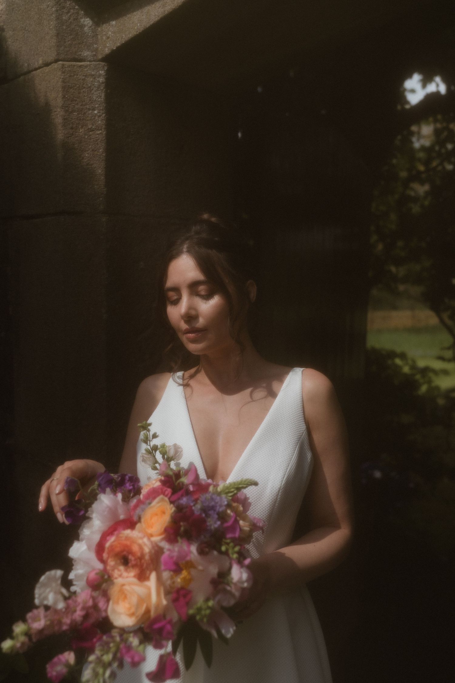 A romantic wedding photo in dark shadows shows a person in a white dress holding a vibrant peach and purple bouquet.